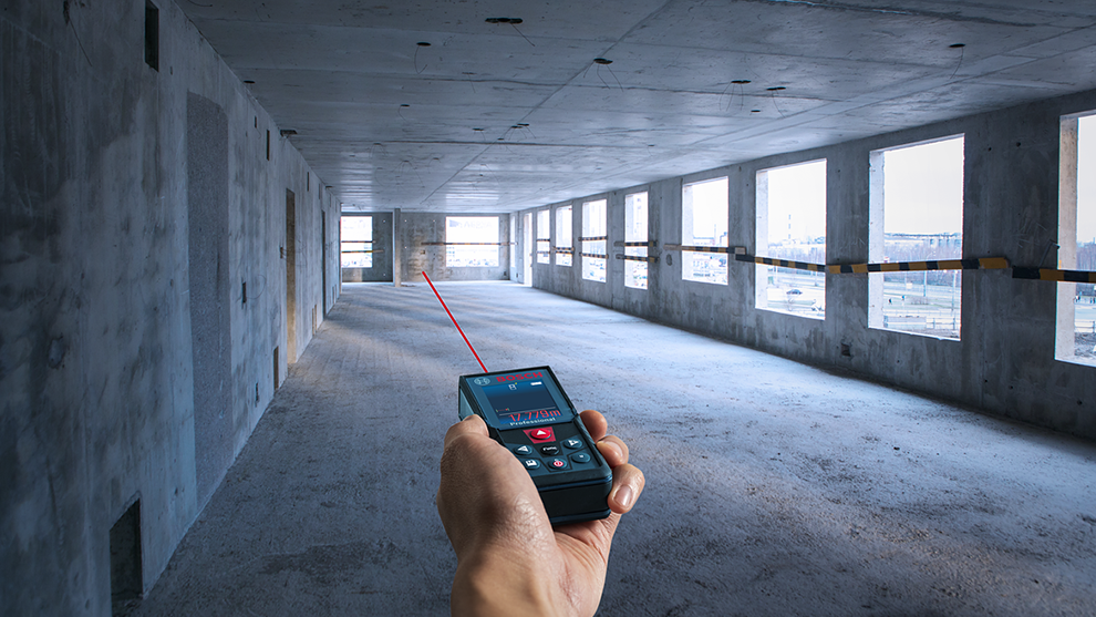 A hand holds a Bosch laser distance measurer in an empty room.