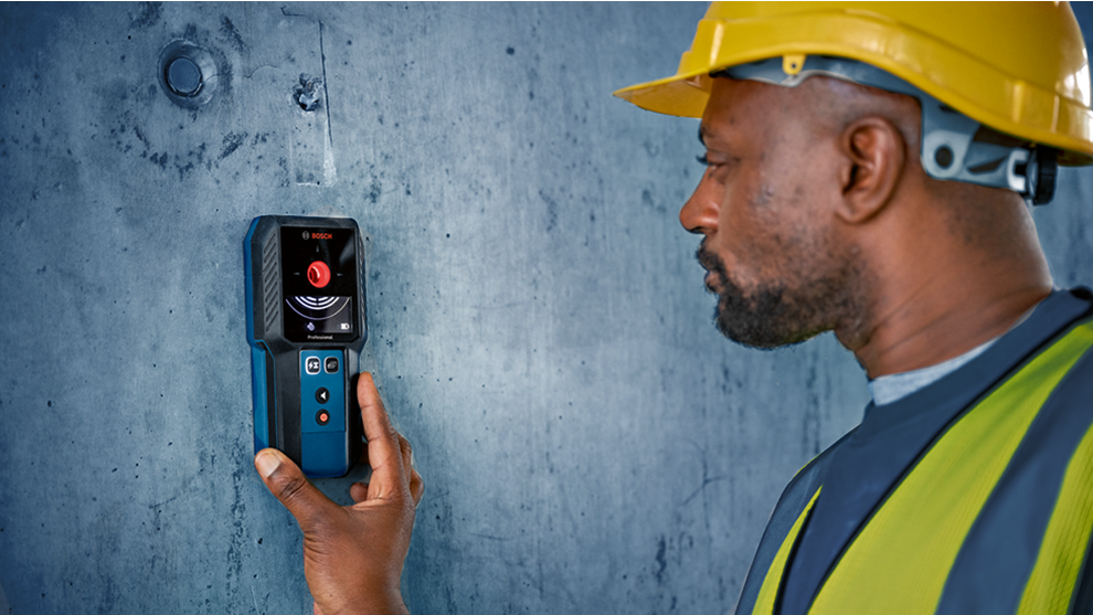 A man with a helmet is using a blue Bosch Professional detector on a wall.