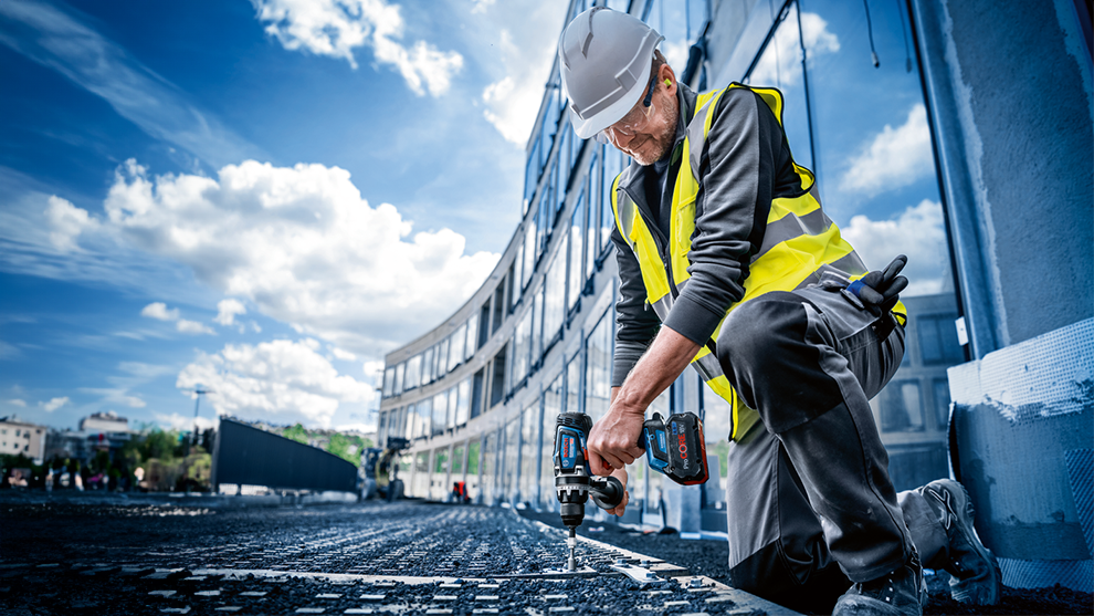 A worker in a yellow vest is using a power screwdriver at a construction site.