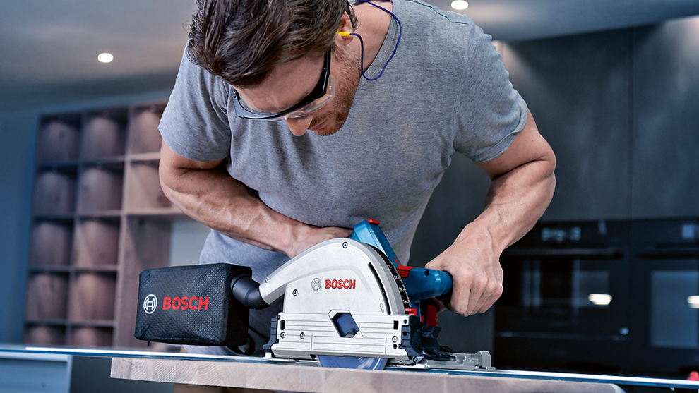 Man with glasses cuts wooden board with Bosch circular saw.