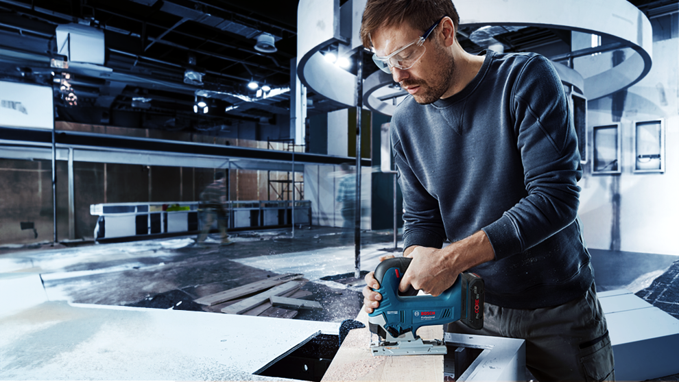 A man wearing safety glasses is using a Bosch Professional jigsaw on wood.