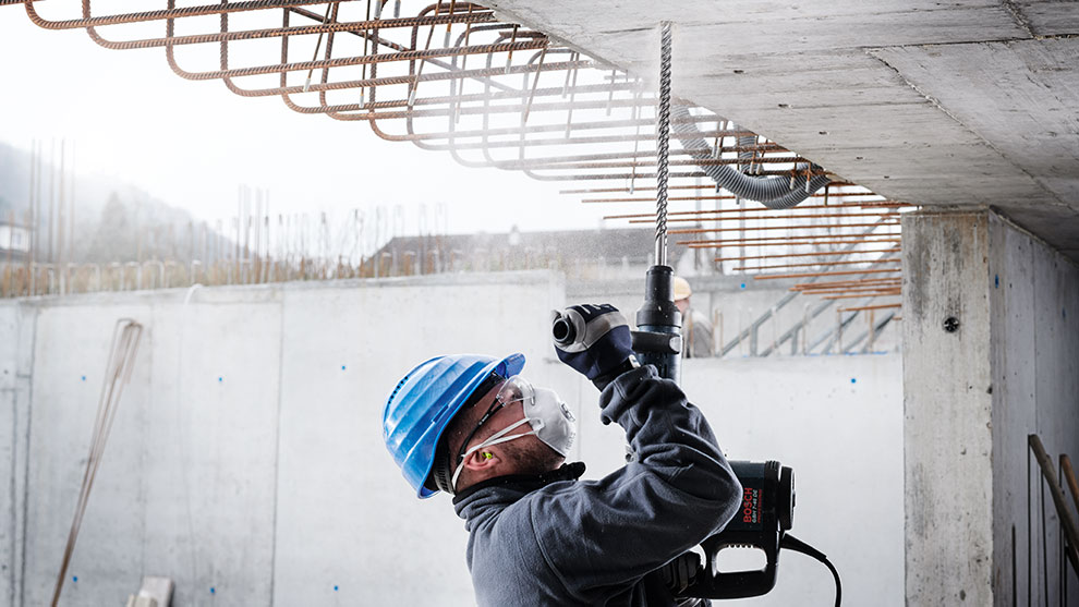 Ein Handwerker mit blauer Helm bohrt in Beton, Sprühnebel sichtbar.