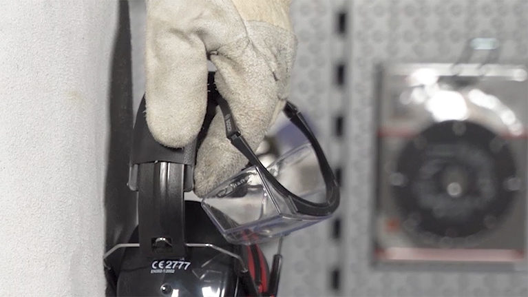 Ruka v bílé rukavici drží ochranné brýle na háčku.