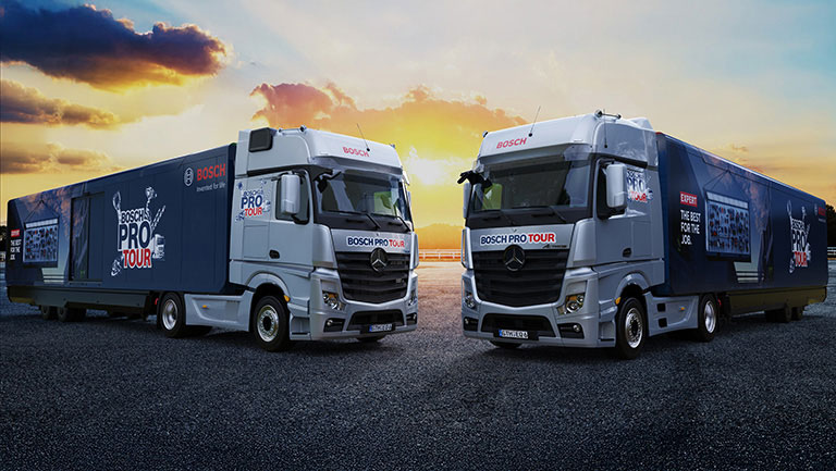 Zwei weiße Lkw stehen auf einer asphaltieren Fläche, mit der Aufschrift "BOSCH PRO TOUR". Der Himmel ist orange und blau.