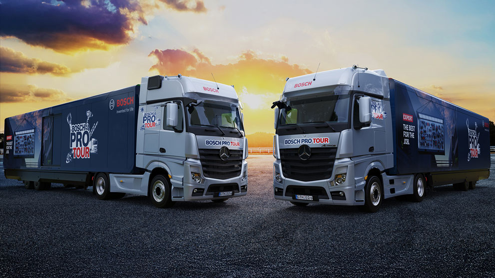 Zwei Mercedes-Lkw stehen auf einer Asphaltfläche, beleuchtet von einem Sonnenuntergang. Beide Lkw haben das Design "BOSCH PRO TOUR".