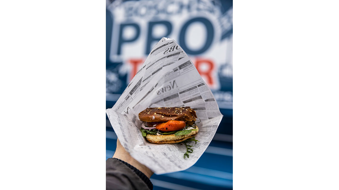 En burger med grøntsager i papir, i baggrunden et blåt banner.