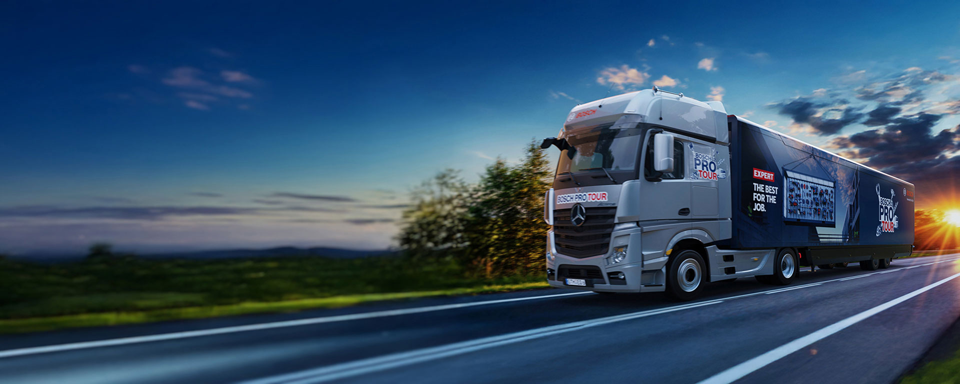 Ein grauer Lkw mit Werbung für "Bosch Pro Tour" fährt auf einer kurvigen Straße bei Sonnenuntergang.