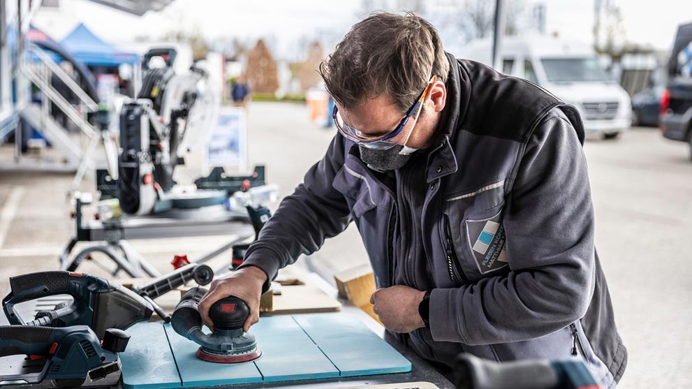 Ein Handwerker mit Schutzbrille schleift ein Holzstück auf einem blau lackierten Tisch, umgeben von Werkzeugen.