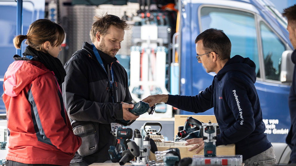 Drei Personen stehen um einen Tisch mit Werkzeugen, beraten sich über Geräte, die Stimmung ist konzentriert.