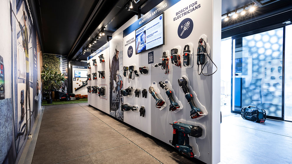 Ein modernes Geschäft mit Werkzeugen. An Wänden hängen verschiedene elektrische Geräte. Ein Bildschirm zeigt Informationen. Der Boden ist aus grauem Stein. Pflanzen stehen in der Ecke, Lichtquellen strahlen gleichmäßig.