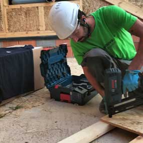 Un artesano con una camisa verde está utilizando un clavador a batería en madera.