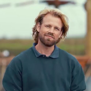 Een man met blond haar en een blauwe trui glimlacht naar de camera.