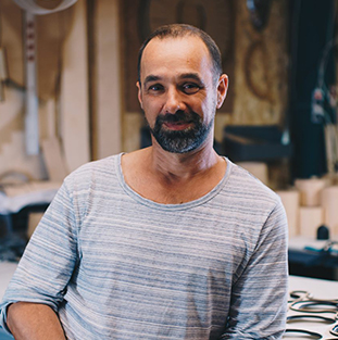 Een man met een baard zit in een werkplaats en glimlacht vriendelijk.