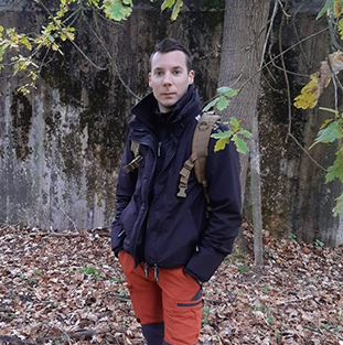 Een man in een zwarte jas en rode broek staat in het bos tussen de bomen.