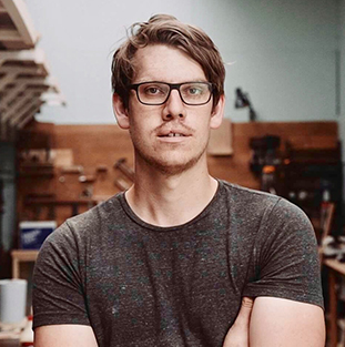 A man with glasses stands in a workshop, arms crossed.