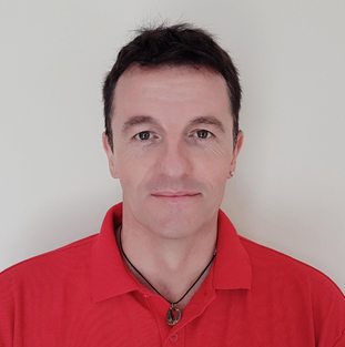 A man in a red shirt stands in front of a neutral background.