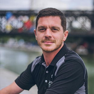 Un homme avec une barbe est assis au bord d'une rivière, avec un pont en arrière-plan.
