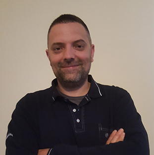 Un homme avec les cheveux courts et une barbe sourit devant un mur lumineux.