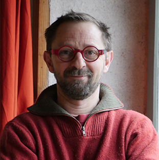 Un homme avec des lunettes et un pull rouge sourit devant une fenêtre.