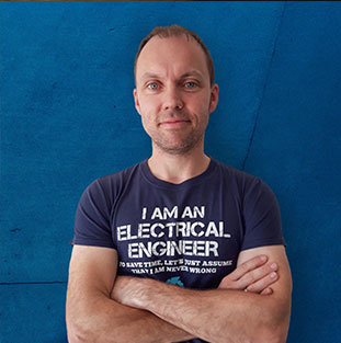 Ein Mann mit T-Shirt steht vor einer blauen Wand mit verschränkten Armen.