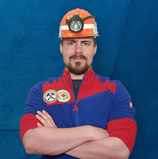 Ein Mann mit Helm und Stirnlampe steht vor blauer Wand, verschränkt die Arme.