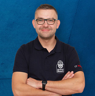 Mann in schwarzem Bosch-Shirt steht vor blauer Wand, lächelt freundlich.