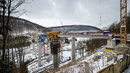Byggnation av en bro över en snötäckt dal med kranar och fordon.