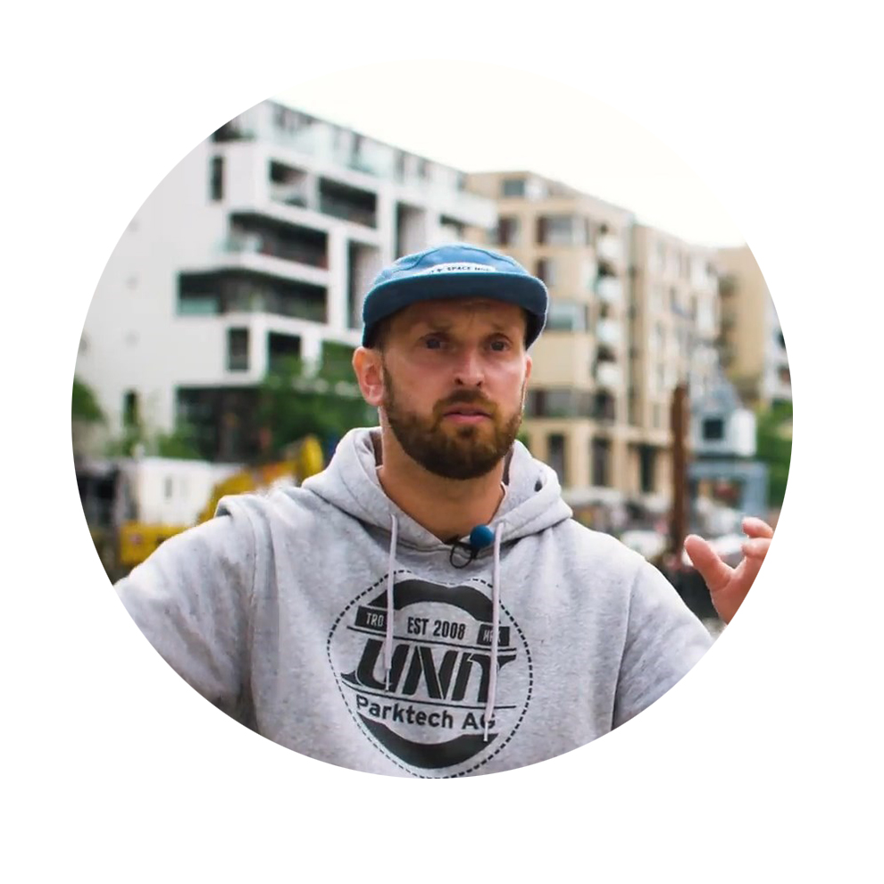 Mees hallis kapuutsiga ja sinise mütsiga räägib modernsete hoonete ees.