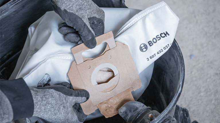 Gloved hands hold a Bosch vacuum cleaner bag over a bucket.