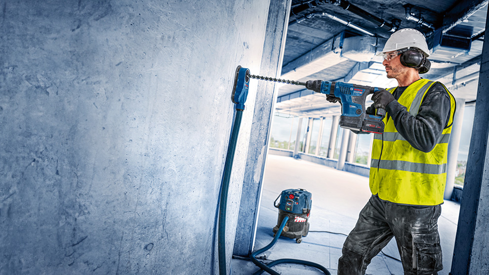A worker wearing a helmet and vest is drilling with a blue Bosch tool.