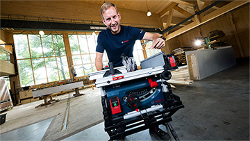 Ein Mann bedient eine Tischsäge in einer hellen Werkstatt mit großen Fenstern.