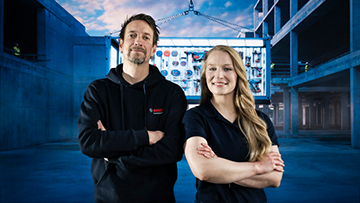 Zwei Personen stehen in einem Bauumfeld, lächelnd, mit Armen vor der Brust verschränkt. Im Hintergrund sieht man eine Baustelle.