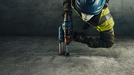 Ein Arbeiter in Sicherheitskleidung nutzt ein Werkzeug auf einem grauen, rauen Boden.