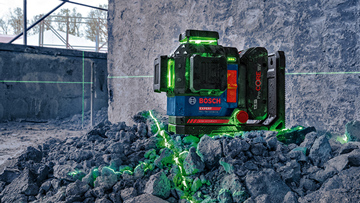 Zelený laserový projektor stojí na šedém štěrku a vyzařuje několik zelených čar na zeď.