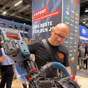 Ein Mann mit Brille und schwarzem T-Shirt hält einen blauen Bosch Professional Akku-Nagler. Im Hintergrund ist ein Messestand mit farbigen Logos und dem Schriftzug "DAS BESTE FÜR DEN JOB" zu sehen. Die Atmosphäre wirkt geschäftig und informativ.