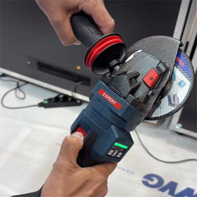 Eine Person hält einen blauen Bosch Professional Winkelschleifer mit einer rot-schwarzen Griffumrandung. Der Schleifer hat eine Scheibe, die bereit ist zum Schneiden. Im Hintergrund ist ein unscharfer Bereich zu sehen, der auf eine Werkstattumgebung hinweist.
