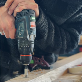 Una persona con un suéter negro sostiene un taladro a batería Bosch Professional, que está perforando una tabla de madera. La máquina es negra con botones verdes y rojos.