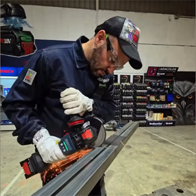 Un hombre con ropa de trabajo y gafas de protección utiliza un amoladora angular Bosch Professional. Chispas vuelan mientras trabaja en un perfil de metal. En el fondo se ven estanterías de herramientas.
