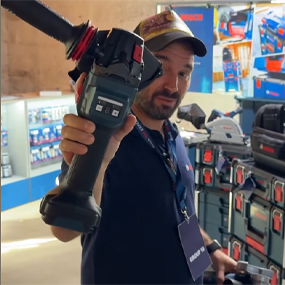 Un hombre con una camisa azul sostiene un destornillador inalámbrico Bosch Professional negro en alto. En el fondo se ven herramientas y cajas de almacenamiento.