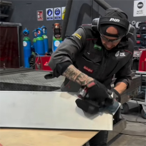 Un trabajador con ropa de trabajo negra está lijando una pieza de metal con una máquina de lijar eléctrica sobre una tabla de madera. En el fondo se pueden ver estaciones de herramientas y botellas de gas de colores. La atmósfera es industrial y profesional.