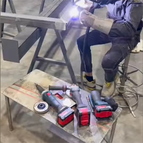 Un trabajador con ropa de trabajo azul está sentado en un taburete y soldando piezas de metal. Delante de él hay varias herramientas Bosch Professional, incluyendo un destornillador inalámbrico, un clavador inalámbrico y una lijadora, todas con acentos rojos. La mesa es de metal y tiene una superficie irregular. En el fondo se pueden ver más perfiles de metal. Un rayo de luz de la máquina de soldadura ilumina la escena, que transmite una atmósfera de trabajo concentrada.