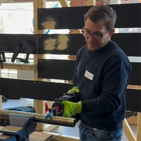 Un homme portant des lunettes et des gants verts utilise un outil sur une table dans un atelier. En arrière-plan, des porte-outils avec d'autres appareils sont visibles.