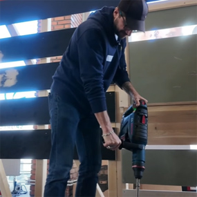 Un homme en vêtements bleus utilise une perceuse sans fil Bosch Professional. Il se tient devant un mur en bois.