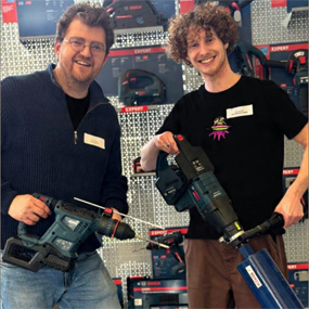 Deux hommes sourient dans un magasin, tenant des outils Bosch Professional : une perceuse et un ciseau.