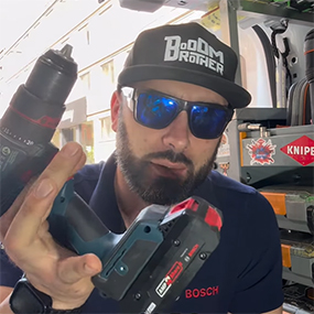 Un uomo con occhiali da sole e un cappellino nero tiene in mano un avvitatore a batteria blu Bosch Professional. Sullo sfondo si vedono strumenti e materiali.