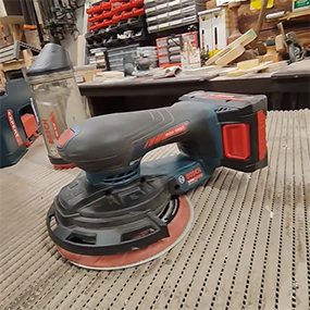 Una levigatrice a batteria nera Bosch Professional con un accento rosso è posata su una superficie di legno, circondata da attrezzi.