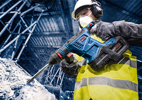 Робітник у попереджувальному одязі використовує синій перфоратор Bosch на будівельному майданчику.