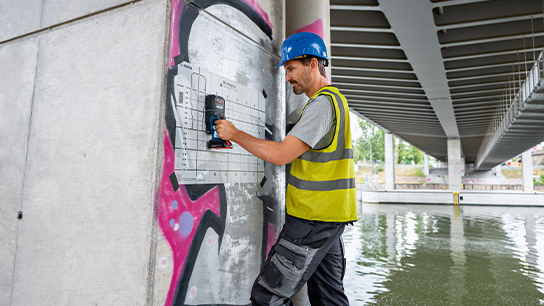 Pracovník s modrou helmou vrtá do betonové zdi pod mostem, obklopen vodou.
