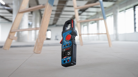 Digitální multimetr stojí na podlaze, dřevěná konstrukce v pozadí, jasná okna.