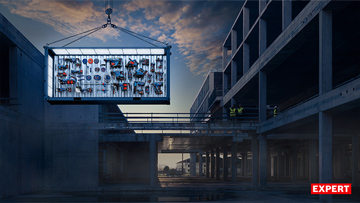 Jeřáb zvedá kontejner s nářadím nad staveniště, obklopené betonovými konstrukcemi.